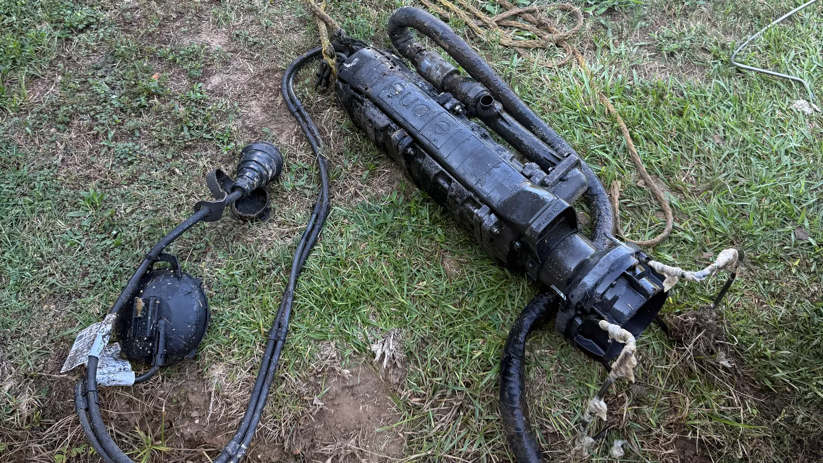 A failed grinder pump pulled out of its basin and laid in the grass beside its float switch and discharge hose, the housing still black with sludge from the tank