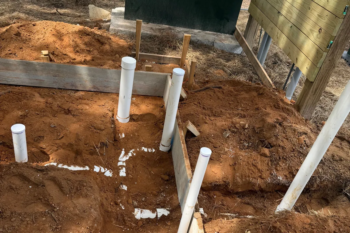 Underground sanitary rough-in on a commercial jobsite, five capped PVC stub-ups standing in red clay along a chalked layout line, with slab form boards set around them and a pier-framed building beyond