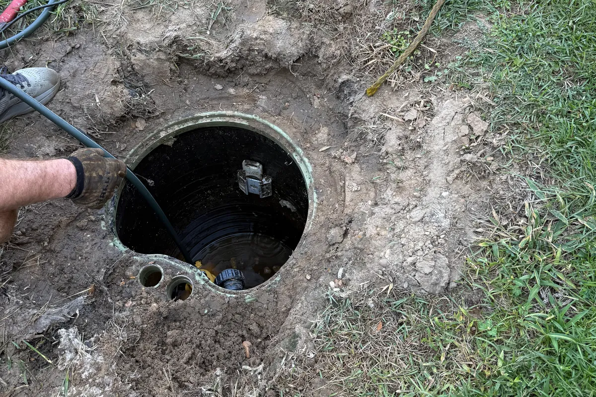 A grinder pump basin opened up in an excavated yard, lid off and set aside, with a plumber rinsing the inside of the tank down with a hose