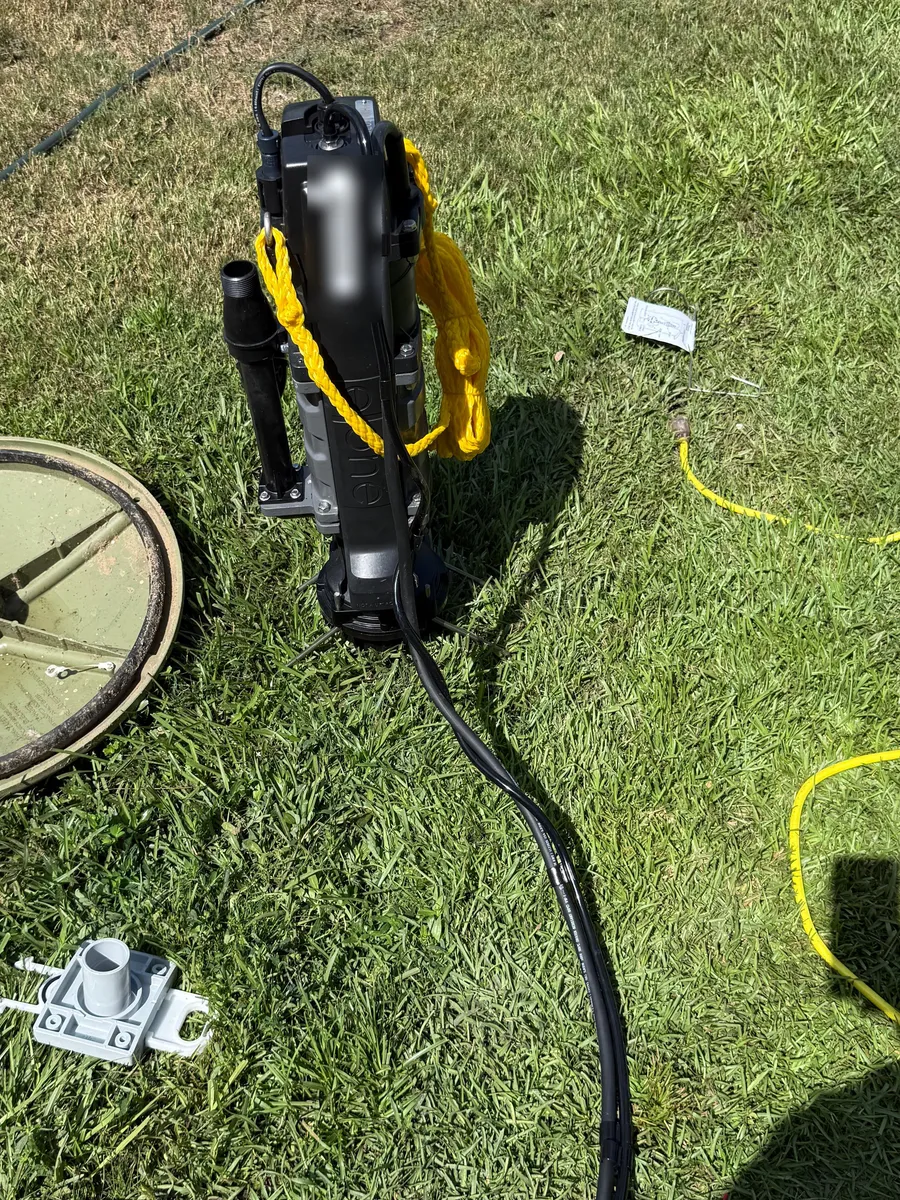 A replacement grinder pump standing in the grass with its lift rope tied off, discharge fitting and float switch laid out beside it, ready to go in the basin