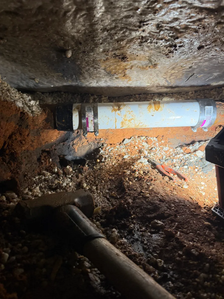 A new white PVC section spliced into an old cast iron drain line with two stainless shielded couplings, set in a hand-dug trench directly beneath a house slab